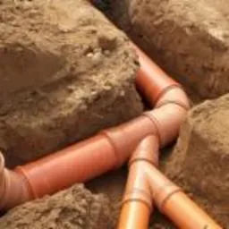 Rury kanalizacyjne z tworzywa sztucznego ułożone w wykopie ziemnym, widoczne złącza i kolanka, świeżo zasypana ziemia.