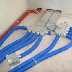 Rozdzielacz wentylacyjny z podłączonymi elastycznymi przewodami w kolorze niebieskim i czerwonymi rurami w tle, widoczne elementy instalacji na surowej podłodze.