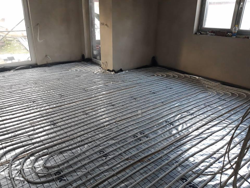 Rozłożone rury systemu grzewczego na podłodze w surowym wnętrzu, widoczne okna i ściany w trakcie wykończenia.