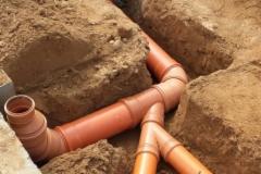 Rury kanalizacyjne z tworzywa sztucznego ułożone w wykopie ziemnym, widoczne złącza i kolanka, świeżo zasypana ziemia.