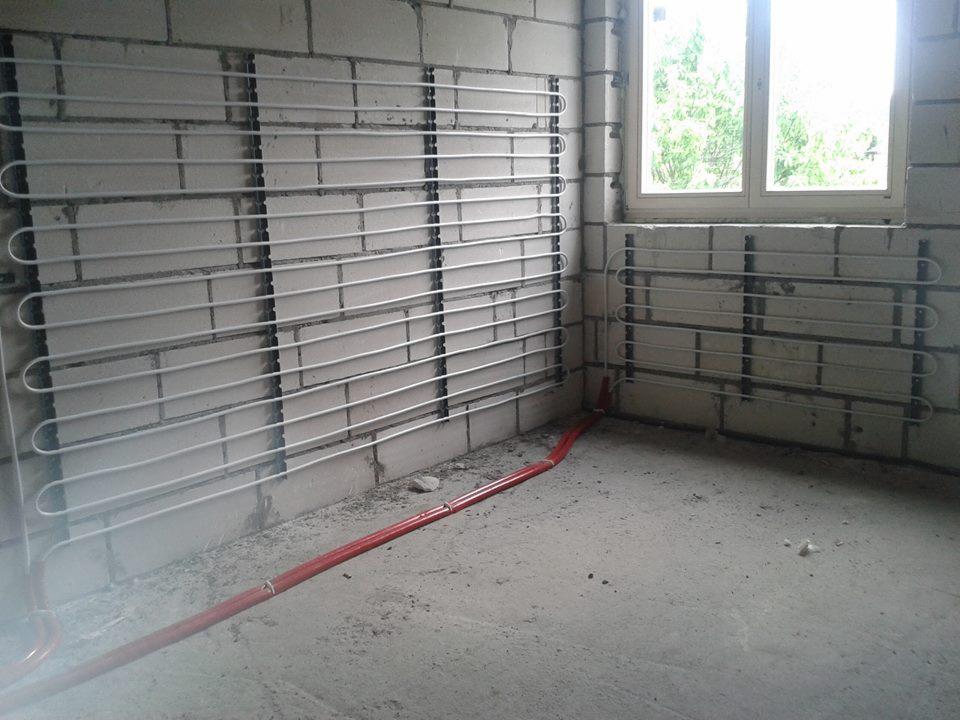 Białe rurki systemu grzewczego zamontowane na surowej ścianie z bloczków betonowych, widoczne czerwone rury doprowadzające, okno w tle.