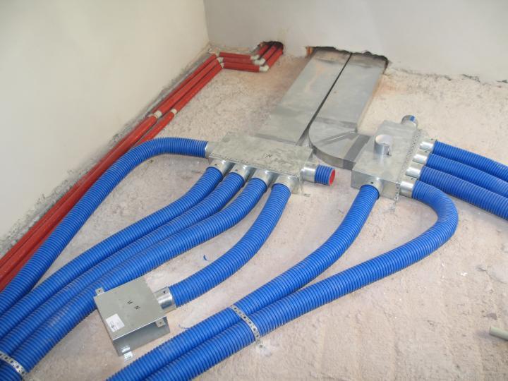 Rozdzielacz wentylacyjny z podłączonymi elastycznymi przewodami w kolorze niebieskim i czerwonymi rurami w tle, widoczne elementy instalacji na surowej podłodze.