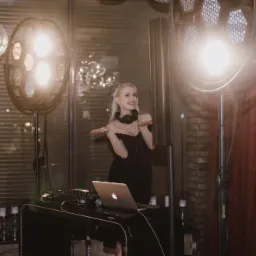 Uśmiechnięta DJ-ka w czarnej sukience i słuchawkach pozuje przy konsolecie DJ-ską z laptopem, otoczona oświetleniem scenicznym i dekoracyjnymi świecami.
