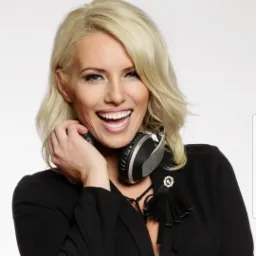 Uśmiechnięta blondynka z słuchawkami na szyi, ubrana w czarną marynarkę z ozdobną broszką i frędzlami, trzymająca słuchawkę przy uchu.