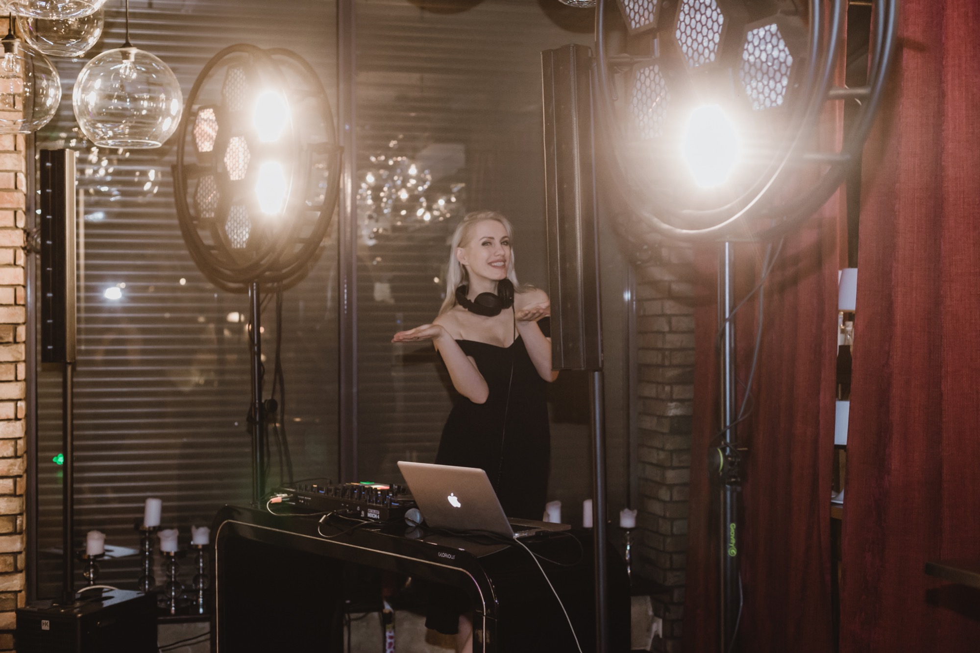 Uśmiechnięta DJ-ka w czarnej sukience i słuchawkach pozuje przy konsolecie DJ-ską z laptopem, otoczona oświetleniem scenicznym i dekoracyjnymi świecami.