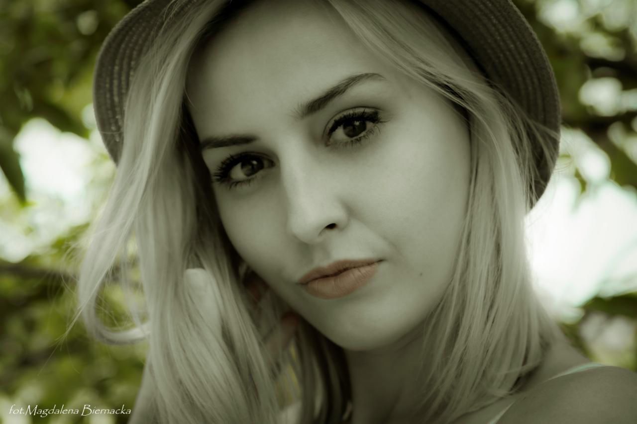 Portret młodej kobiety z blond włosami i kapeluszem na tle zieleni, wykonany w sepii z delikatnym uśmiechem.