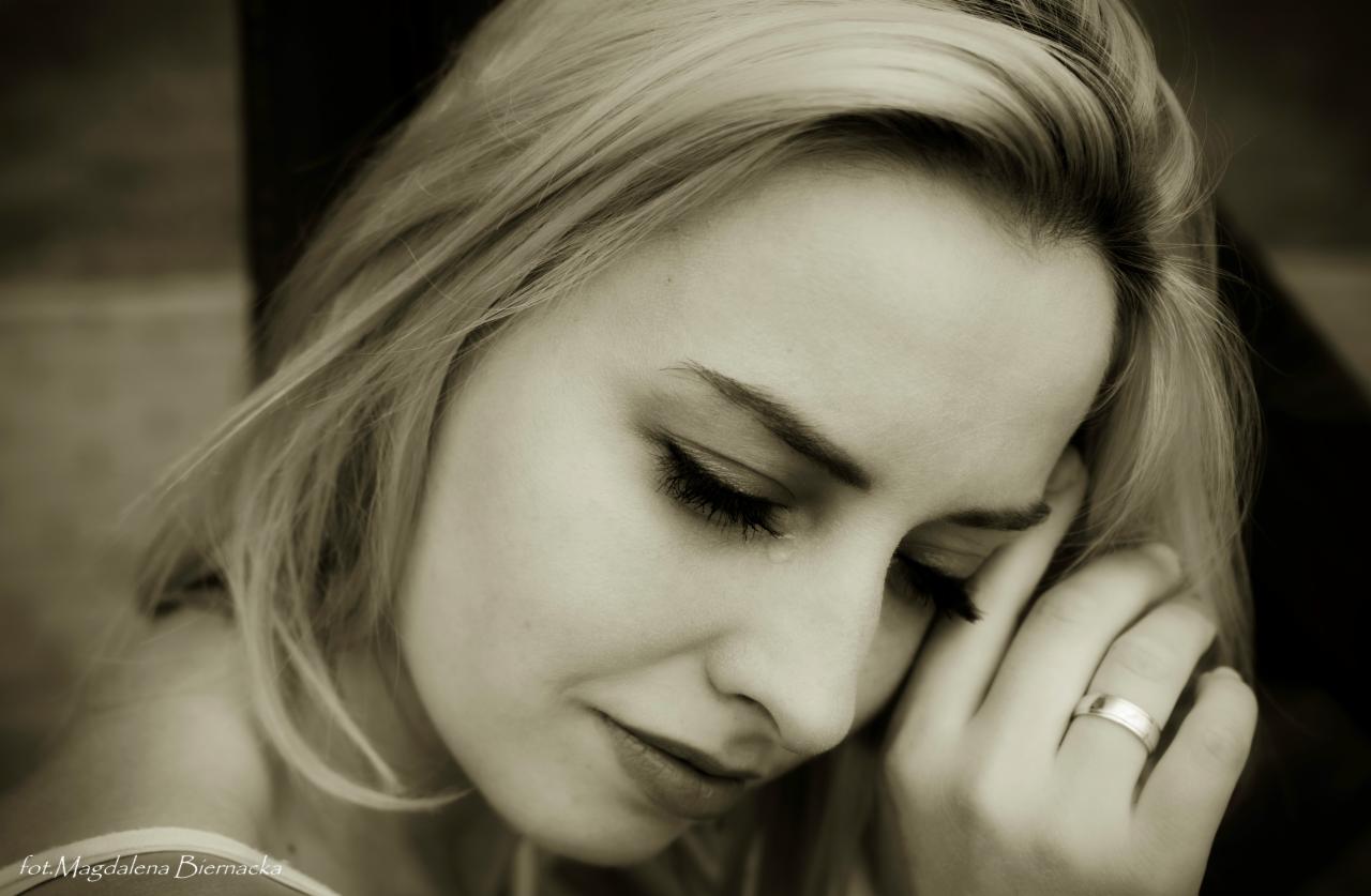 Portret kobiety z blond włosami, zamyślona, oparta policzkiem na dłoni z widocznym pierścionkiem.