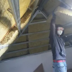Pracownik w masce i okularach ochronnych montuje wełnę mineralną w konstrukcji metalowej poddasza, widoczne elementy izolacji termicznej i konstrukcja szkieletowa dachu.