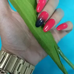 Dłoń z pomalowanymi paznokciami w odcieniach czerwieni i czerni (ombre), trzymająca zielony łodygę sztucznego kwiatu na tle błękitu, z widocznym złotym zegarkiem Michael Kors na nadgarstku.