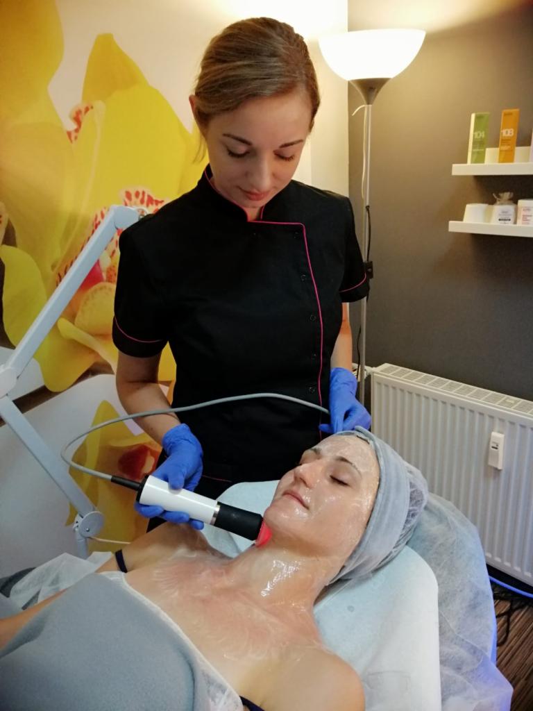 Kosmetolog w niebieskich rękawiczkach wykonuje zabieg na twarzy klientki za pomocą urządzenia emitującego czerwone światło, na tle ściany z motywem kwiatowym i półek z kosmetykami.