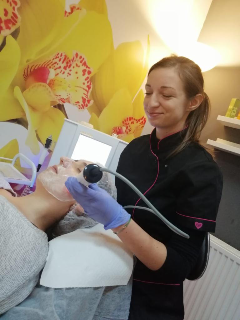 Kosmetolog w czarnym uniformie z różowym obszyciem wykonuje zabieg na twarz klientki za pomocą urządzenia z okrągłą głowicą, na ścianie widoczna tapeta z motywem żółtych orchidei.