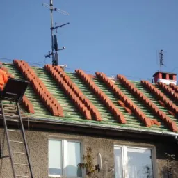 Montaż dachówki ceramicznej na zielonej membranie dachowej; pracownik w pomarańczowej kurtce na drabinie z platformą roboczą, widoczne anteny satelitarne na dachu budynku.