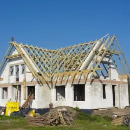 Surowy stan domu jednorodzinnego w trakcie budowy, widoczne bloczki Ytong, drewniana konstrukcja dachu i niezadaszone okna.