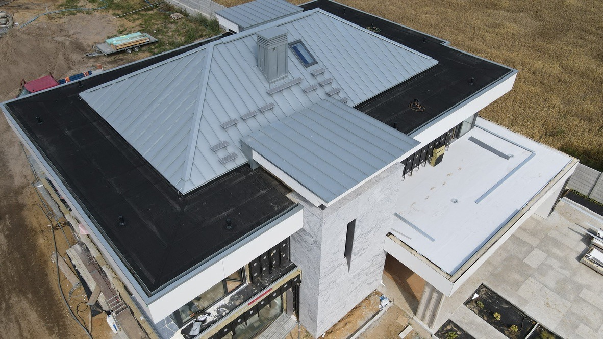 Nowoczesny dom z szarą, metalową dachówką i płaskim dachem pokrytym czarną membraną. Widok z góry ukazuje detale wykończenia i elementy architektoniczne.