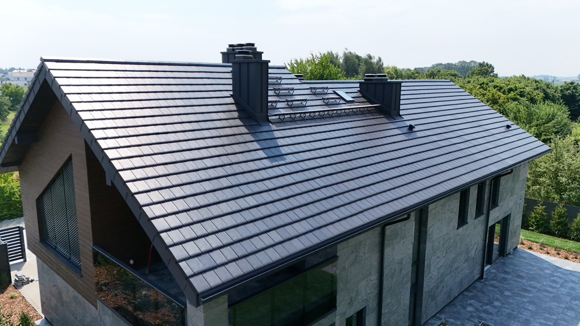 Nowoczesny dom jednorodzinny z ciemną, płaską dachówką zakładkową, widok z góry na dach, kominy i okna dachowe. Tło stanowią drzewa i błękitne niebo.