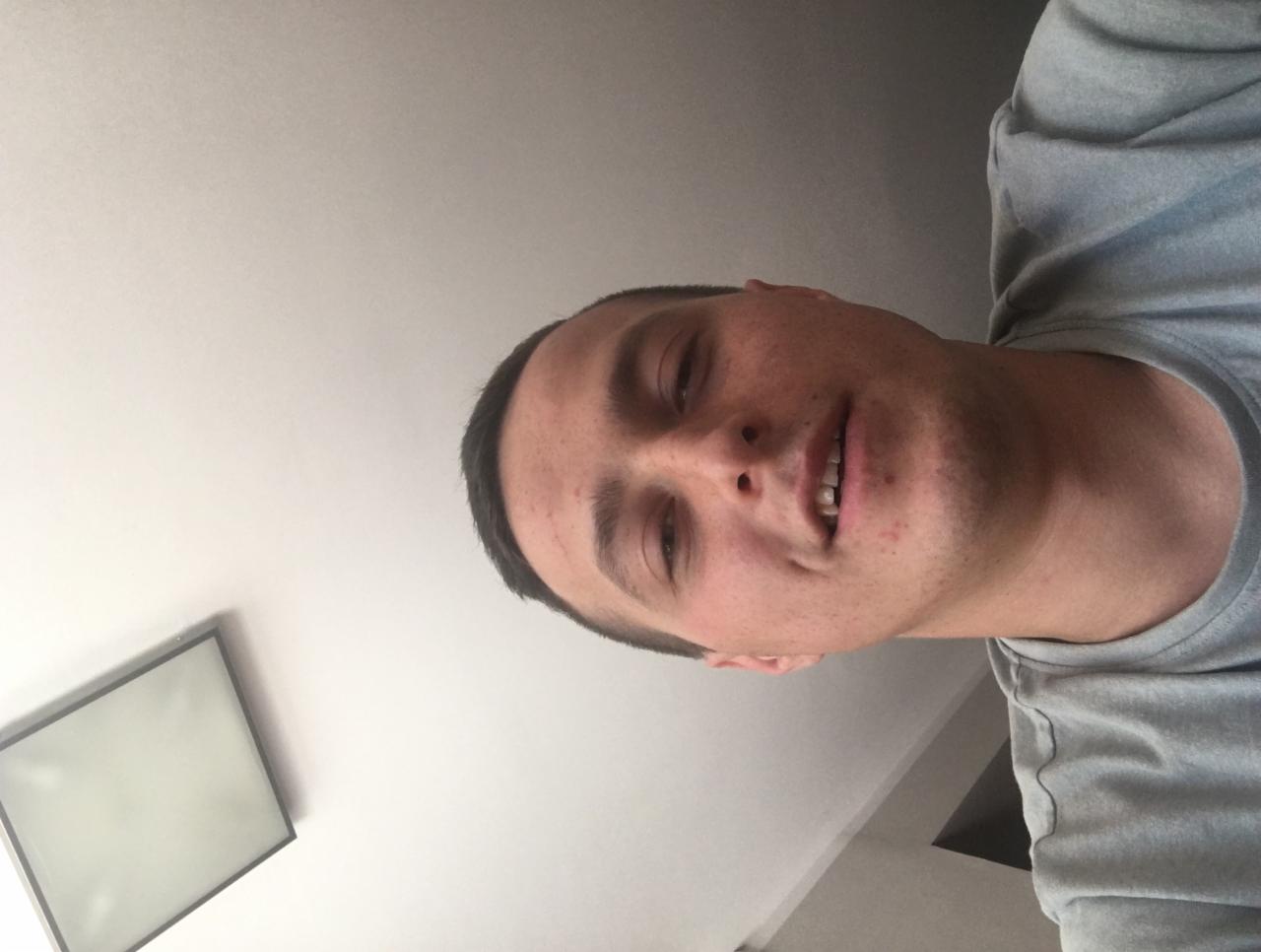 Portret mężczyzny z krótkimi włosami, ujęcie z dołu, widoczny fragment sufitu i lampy. Uśmiechnięty, ubrany w szarą koszulkę.