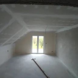 Poddasze po montażu płyt gipsowo-kartonowych, widoczne łączenia na suficie i ścianach, niepomalowane, z drewnianą łatą na podłodze i oknem balkonowym w tle.