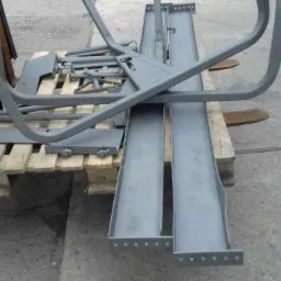 Metalowe elementy konstrukcyjne, świeżo pomalowane na szaro, ułożone na drewnianej palecie na betonowym podłożu. Widoczne ślady niedoskonałości w malowaniu oraz niewielkie zarysowania.