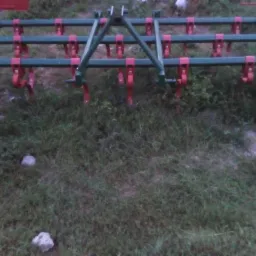 Kultywator rolniczy z czerwonymi zębami i zielonymi kołami, stojący na trawie.