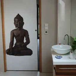 Nowoczesna łazienka z umywalką nablatową, minimalistyczną armaturą i dużym lustrem; na drzwiach naklejka z wizerunkiem Buddy, dodająca wnętrzu elementu medytacyjnego spokoju.