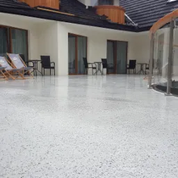 Jasnoszara, błyszcząca posadzka na tarasie z widocznymi detalami faktury, z trzema leżakami z logo i krzesłami stolikowymi w tle oraz szklaną balustradą po prawej stronie.