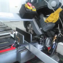 Piła taśmowa do metalu w trakcie cięcia profilu, widoczne elementy sterujące, oświetlenie robocze i fragment samochodu w tle.