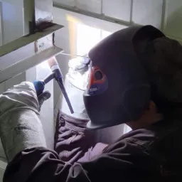 Pracownik w masce spawalniczej łączy metalowe elementy konstrukcyjne za pomocą spawarki TIG w jasnym pomieszczeniu.