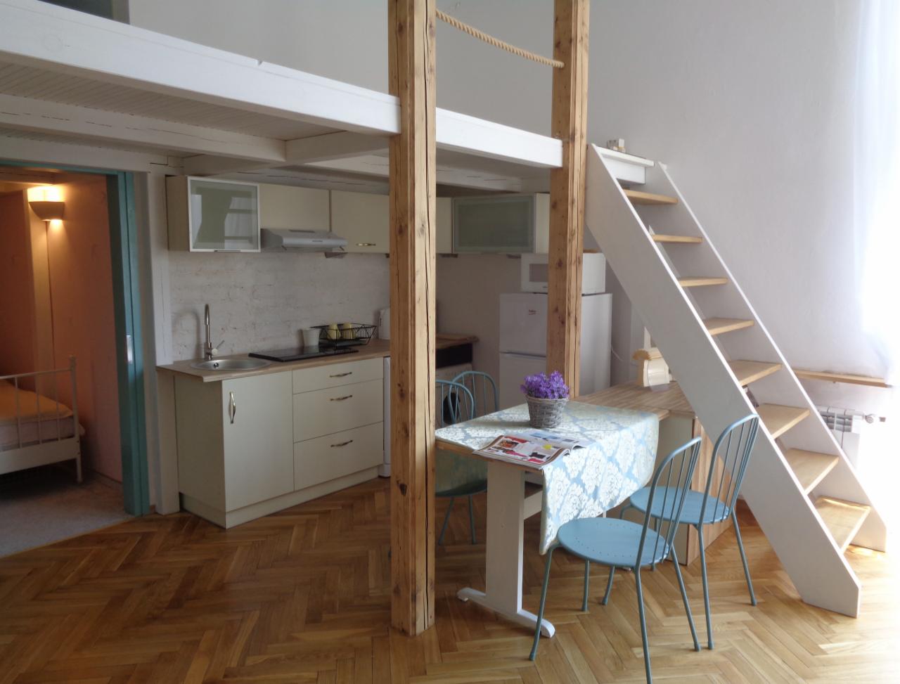 Jasne wnętrze z antresolą nad kuchnią, drewnianymi słupami i schodami, parkiet w jodełkę, stół z kwiatami i krzesłami.