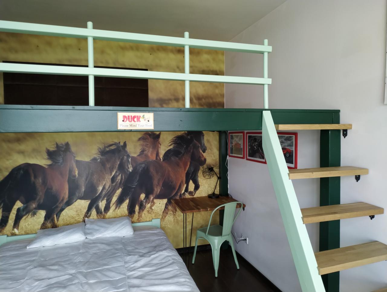 Pokój z łóżkiem na antresoli, pomalowanym na zielono, z drewnianymi schodami i biurkiem pod spodem, tapeta z motywem biegnących koni na ścianie za łóżkiem.