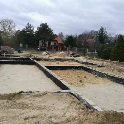 Przygotowanie fundamentów pod budynek jednorodzinny: wykopane i częściowo zalane piaskiem zbrojone ławy fundamentowe, widoczne elementy izolacji pionowej, teren budowy z hałdami ziemi w tle.