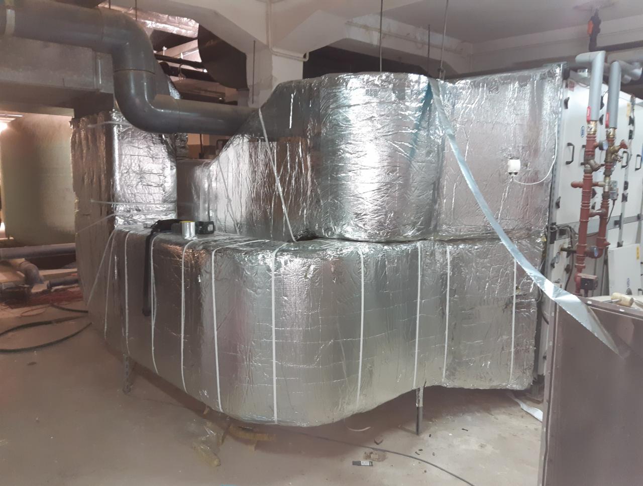 Izolacja termiczna instalacji przemysłowej, owinięta folią aluminiową i zabezpieczona taśmą, widoczne rury i elementy armatury w tle.