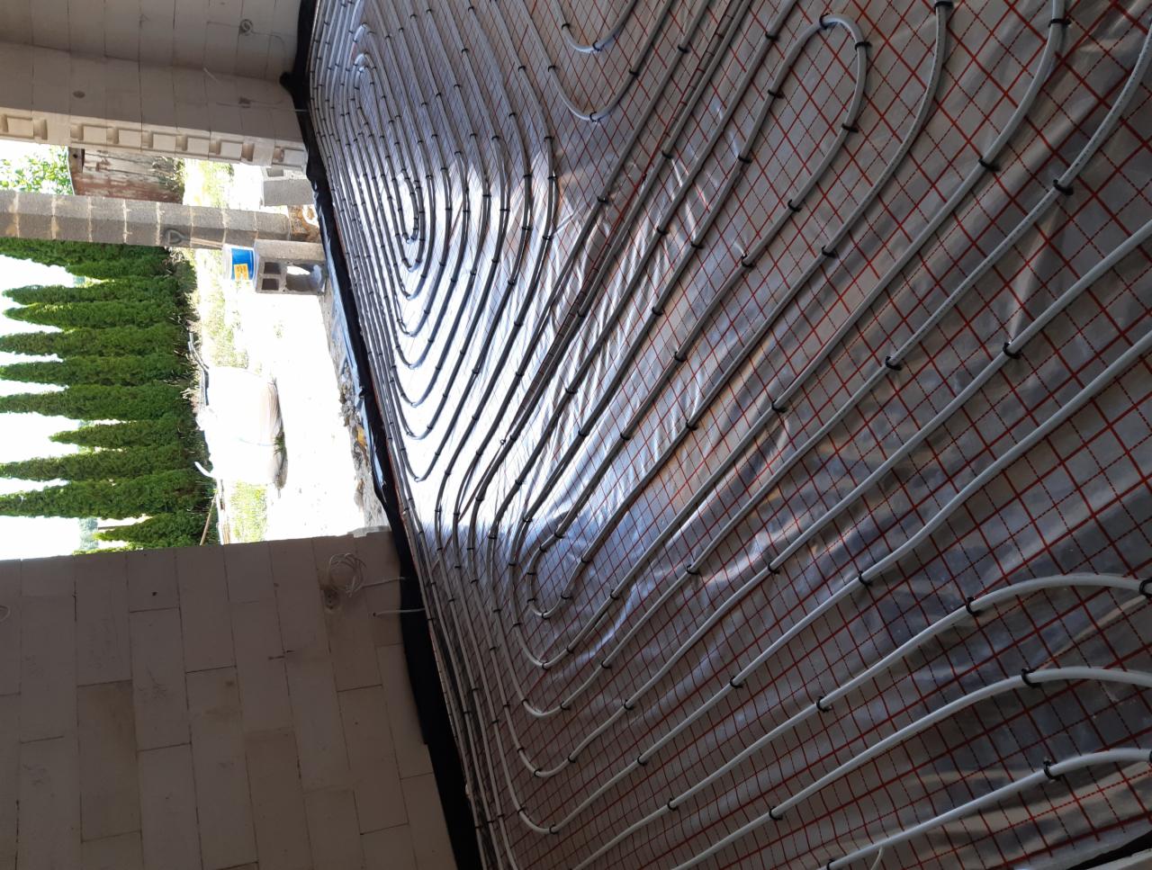 Białe rury systemu ogrzewania podłogowego ułożone na srebrnej folii z czerwoną siatką w trakcie montażu, widok z góry z perspektywy narożnika pomieszczenia, z widokiem na zewnątrz przez otwór okienny.