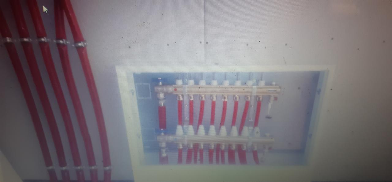 Rozdzielacz ogrzewania podłogowego w białej szafce, widoczne czerwone rury doprowadzające i odprowadzające wodę, instalacja w trakcie montażu.