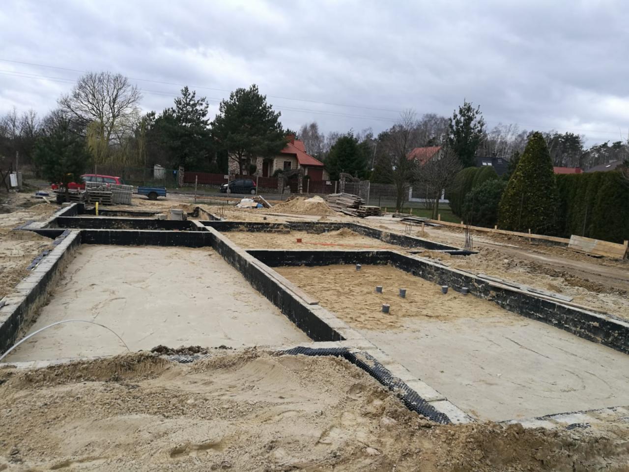 Przygotowanie fundamentów pod budynek jednorodzinny: wykopane i częściowo zalane piaskiem zbrojone ławy fundamentowe, widoczne elementy izolacji pionowej, teren budowy z hałdami ziemi w tle.