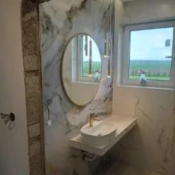 Łazienka w trakcie remontu: marmurowe płytki na ścianie, owalne lustro w złotej ramie, biała umywalka nablatowa z baterią w kolorze złota, wiszące lampy tuby w kolorze złota, okno z widokiem...