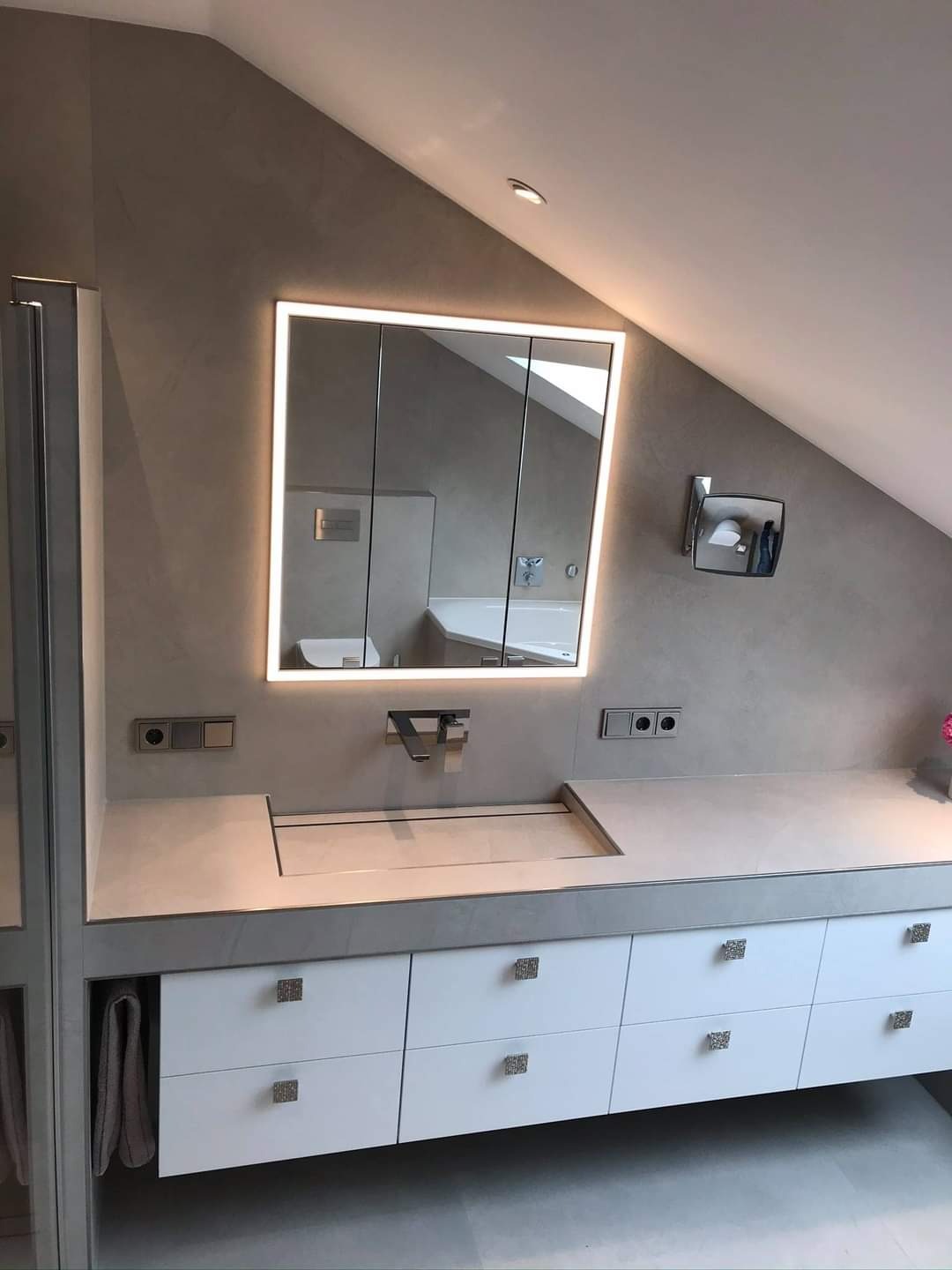 Nowoczesna łazienka na poddaszu ze skośnym sufitem, oświetlonym lustrem szafkowym, białą szafką pod umywalką z kwadratowymi uchwytami i ukrytą umywalką pod blatem.