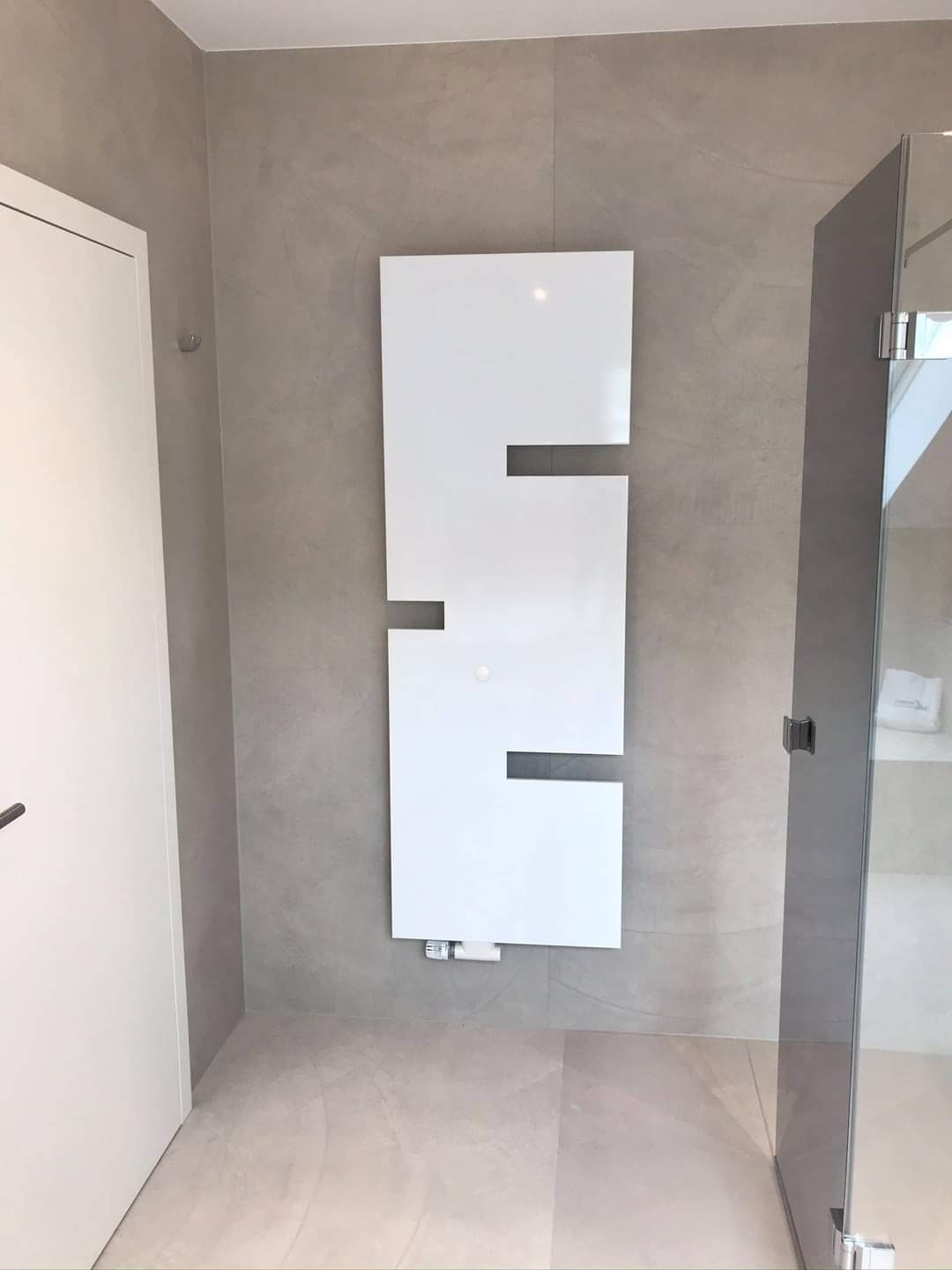 Nowoczesna łazienka z białym, dekoracyjnym grzejnikiem panelowym na szarej ścianie, widoczna kabina prysznicowa ze szklanymi drzwiami i białe drzwi wejściowe.