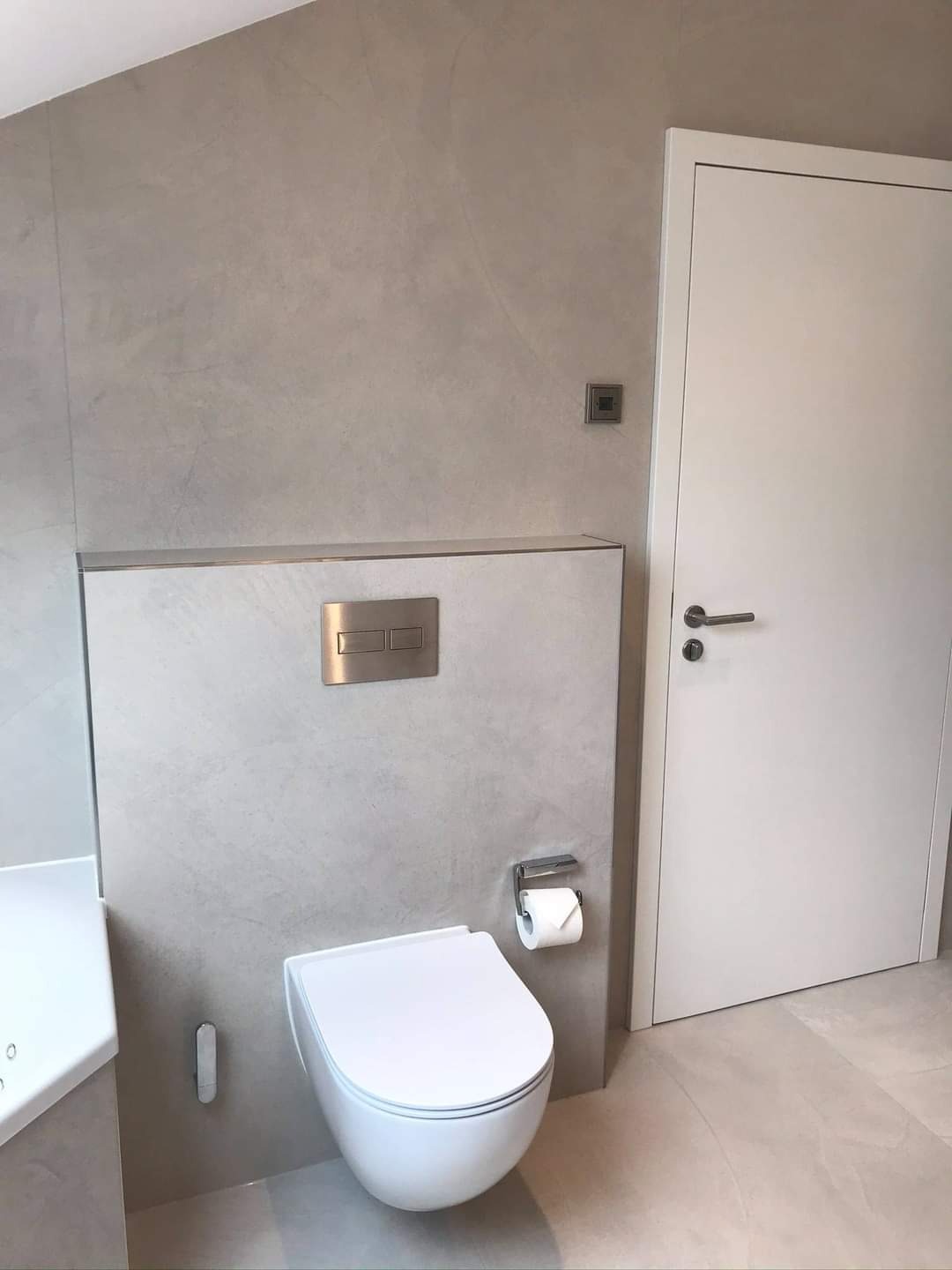 Nowoczesna, minimalistyczna toaleta podwieszana z chromowanymi przyciskami spłukiwania i uchwytem na papier toaletowy, na tle ściany z efektem dekoracyjnym i białych drzwi.