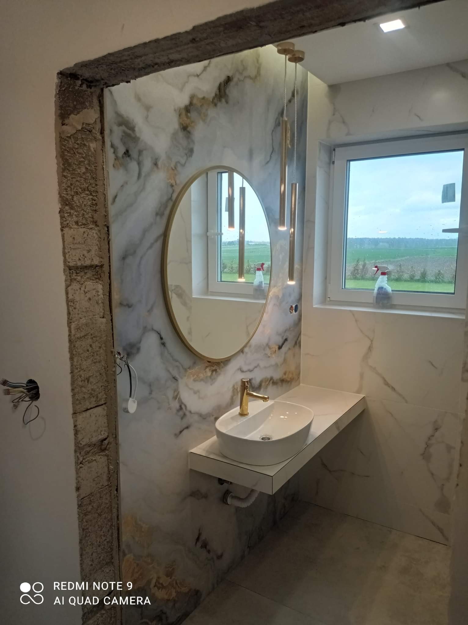 Łazienka w trakcie remontu: marmurowe płytki na ścianie, owalne lustro w złotej ramie, biała umywalka nablatowa z baterią w kolorze złota, wiszące lampy tuby w kolorze złota, okno z widokiem...
