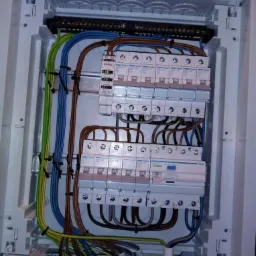 Rozdzielnica elektryczna z zainstalowanymi bezpiecznikami Hager, okablowaniem w kolorach niebieskim, brązowym, zielono-żółtym, widoczne szyny DIN i listwy zaciskowe.