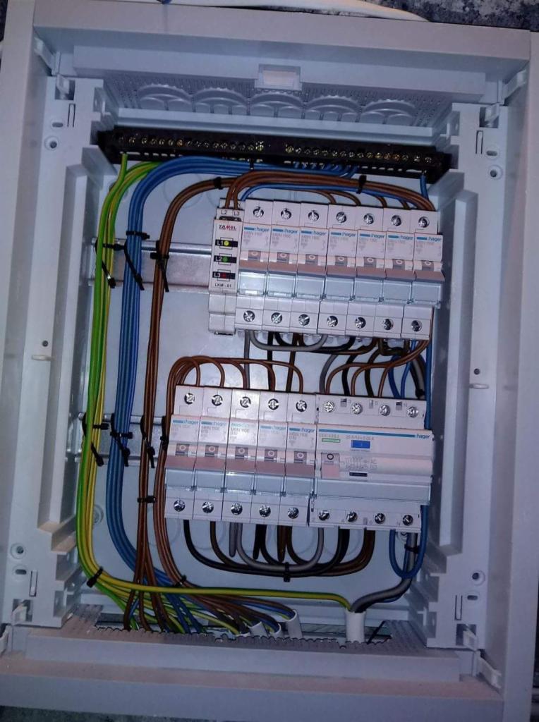 Rozdzielnica elektryczna z zainstalowanymi bezpiecznikami Hager, okablowaniem w kolorach niebieskim, brązowym, zielono-żółtym, widoczne szyny DIN i listwy zaciskowe.