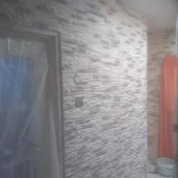 Ściana wykończona dekoracyjną okładziną imitującą kamień, z widocznym włącznikiem światła i zabezpieczonym folią wejściem do pomieszczenia, w tle fragment zasłony i wiaderko.