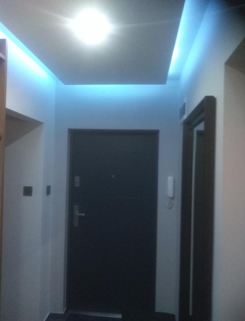 Nowoczesny przedpokój z podwieszanym sufitem i niebieskim oświetleniem LED, ciemne drzwi wejściowe.