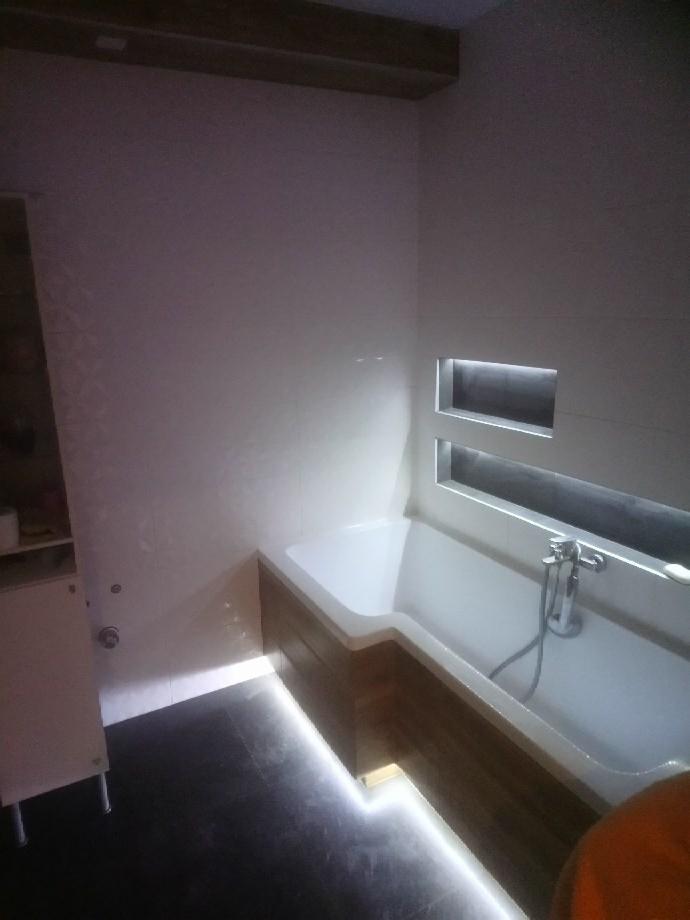 Nowoczesna łazienka z podświetlaną wanną obudowaną drewnem, czarną podłogą i wnękami z oświetleniem LED w ścianie.