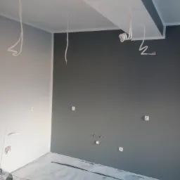 Pok&oacute;j w trakcie remontu: ściany pomalowane na dwa odcienie szarości, widoczne kable wystające z sufitu i ściany, zabezpieczona podłoga, narzędzia malarskie i wiertarka na podłodze.