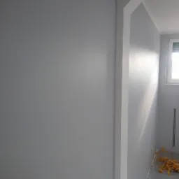 Świeżo pomalowana na szaro ściana w Luszowicach, z widocznymi śladami taśmy malarskiej na podłodze i białym wykończeniem framugi drzwi.