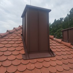 Budmar - Ceglasty dach z ceramiczną dachówką karpiówką i brązowym, metalowym kominem. Widoczne drzewa w tle i pochmurne niebo. Komin z blachy na dachu.