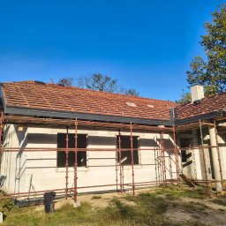 Budmar - Wznoszony dom z białego pustaka, z częściowo ułożoną czerwoną dachówką na łatach. Widoczne rusztowania i fragmenty drzew na tle błękitnego nieba.