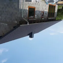 Surowy budynek z szarego betonu komórkowego z zamontowanymi brązowymi oknami i ciemnym dachem z rynnami, na tle błękitnego nieba.