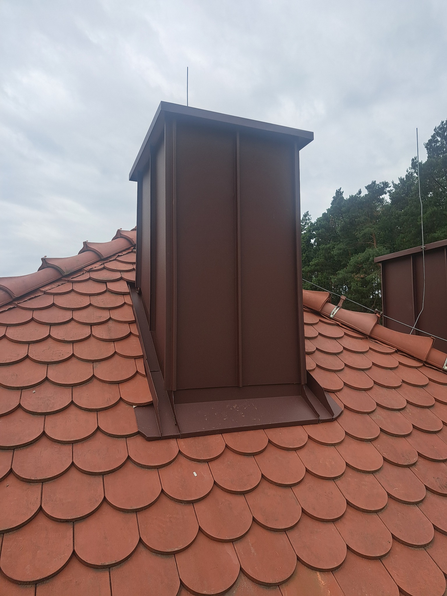Ceglasty dach z ceramiczną dachówką karpiówką i brązowym, metalowym kominem. Widoczne drzewa w tle i pochmurne niebo. Komin z blachy na dachu.
